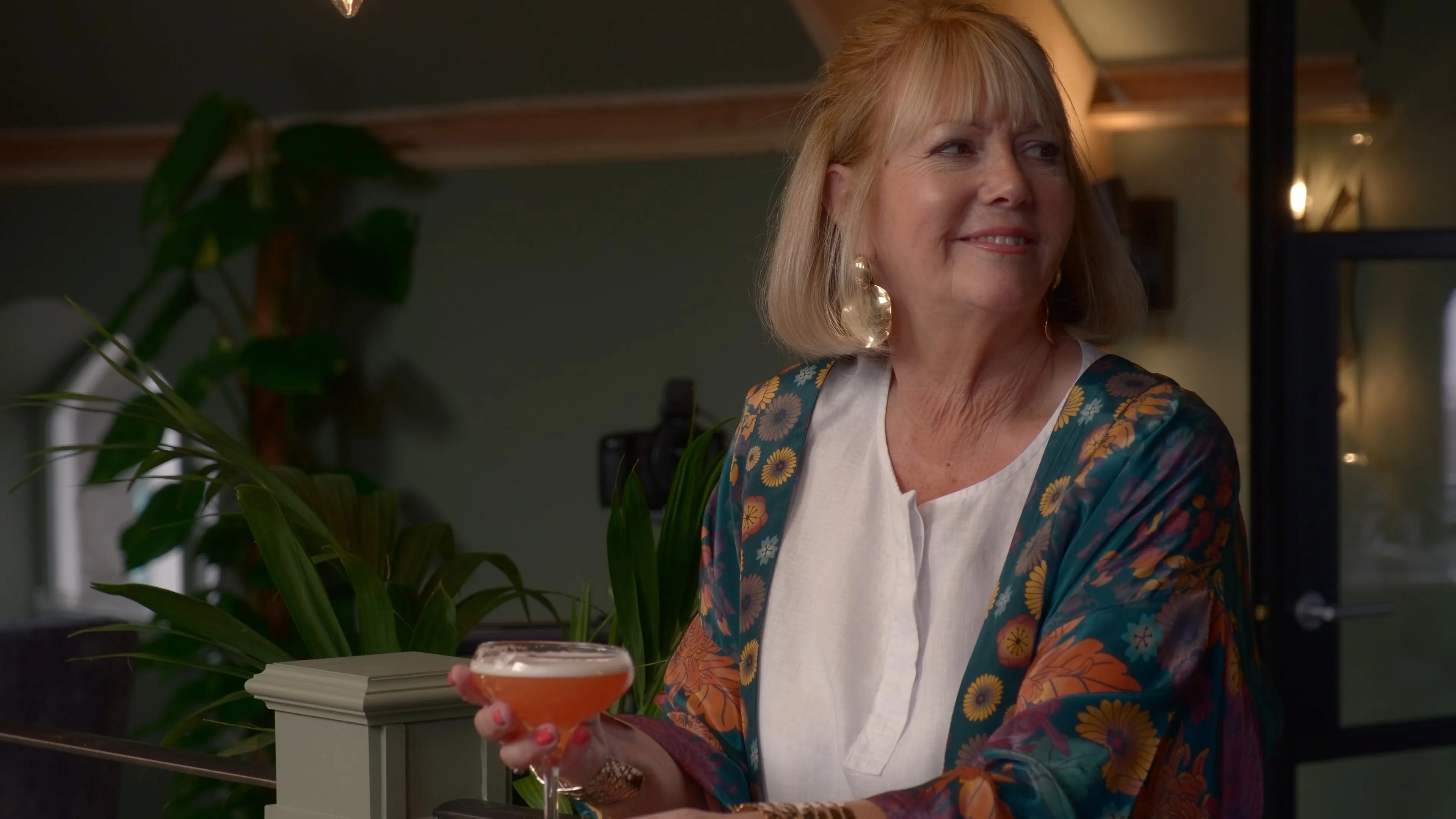 
A smiling woman with blonde hair holding a pink cocktail glass in the warm, plant-filled interior of Kella's restaurant, Port St Mary, Isle of Man. She is wearing a colourful floral kimono-style jacket.